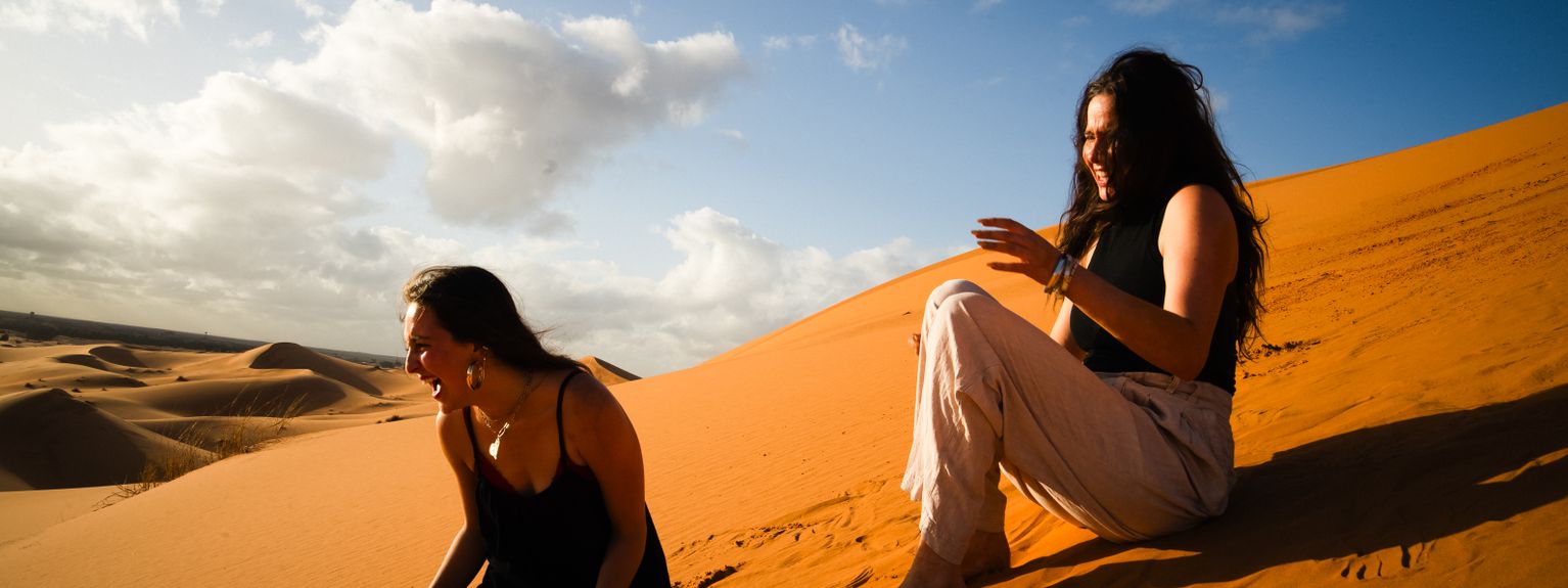Two people sitting and laughing on a sunlit sand dune, with a blue sky and scattered clouds in the background.