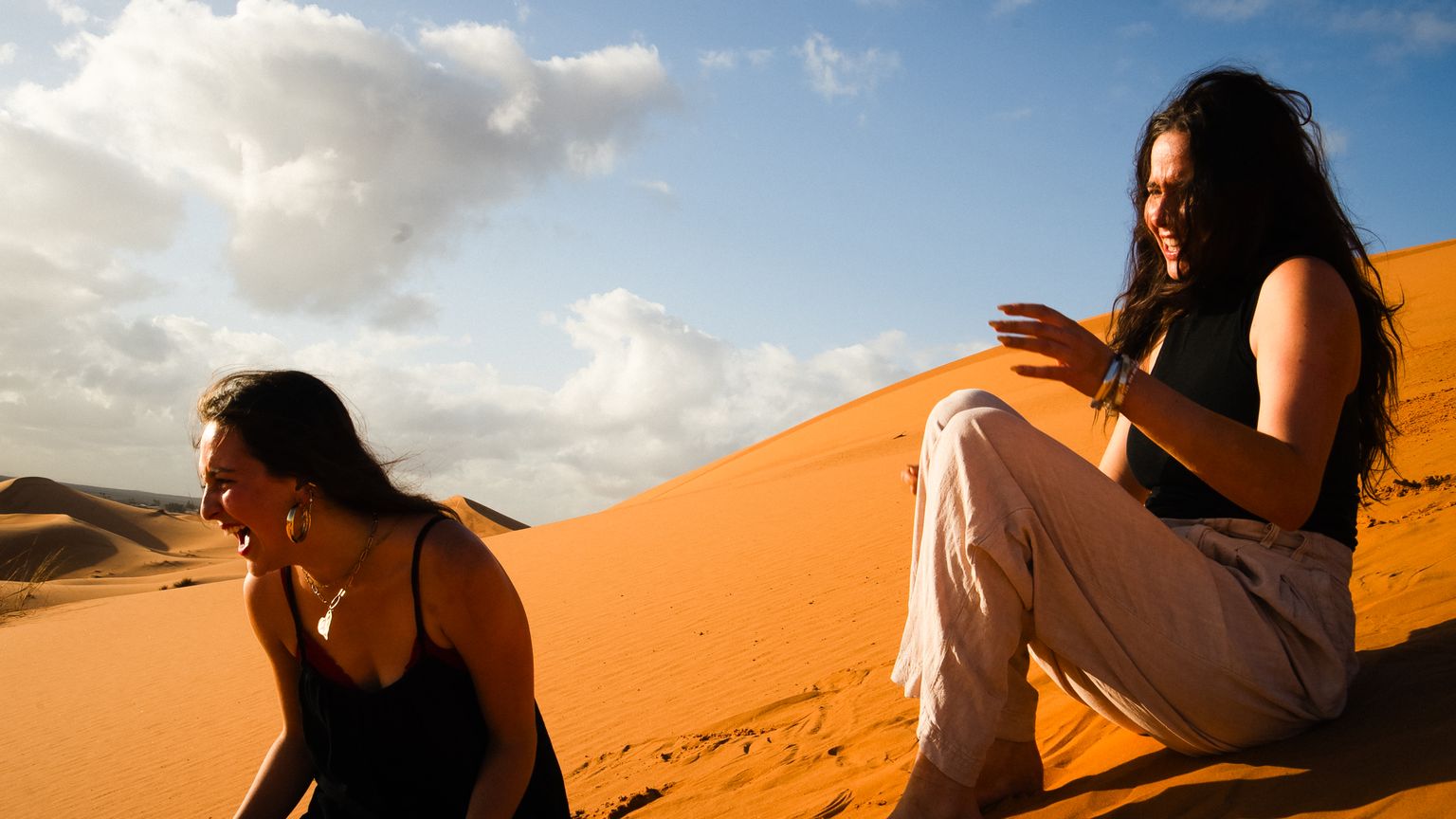 Two people sitting and laughing on a sunlit sand dune, with a blue sky and scattered clouds in the background.