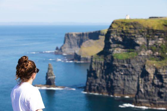 Person with sunglasses admires the scenic view of the Cliffs of Moher, with rugged cliffs and blue ocean under a clear sky.