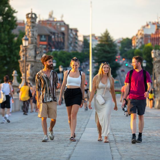 Four people happily walking across a sunlit bridge in a city, with historic stone railings and a bustling backdrop.