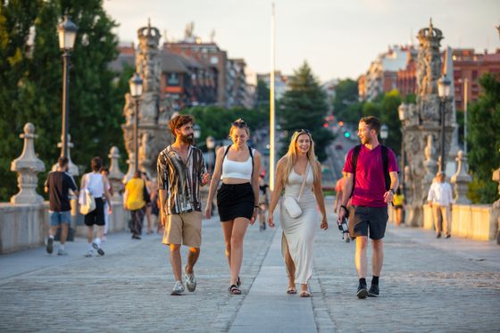 Four people walking and smiling on a cobblestone bridge lined with lamps, surrounded by blurred pedestrians and greenery in the background.