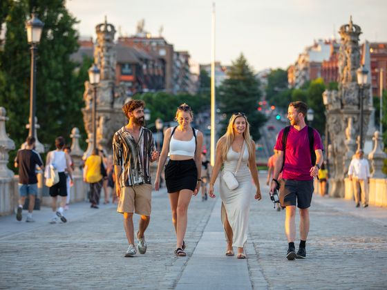 Four people walking and smiling on a cobblestone bridge lined with lamps, surrounded by blurred pedestrians and greenery in the background.
