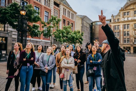 A tour guide gestures upward while a group of people attentively look in the same direction, standing in a city square with historic buildings.