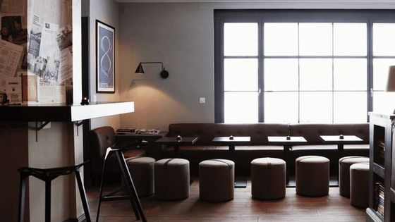 Minimalist cafe interior with modern furniture, a row of stools by a wall bench, wooden flooring, and a large window offering natural light.