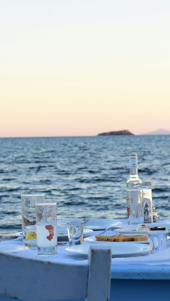 Outdoor dining setup by the sea at sunset, featuring empty plates, glasses, a bottle of water, and bread on a wooden table.