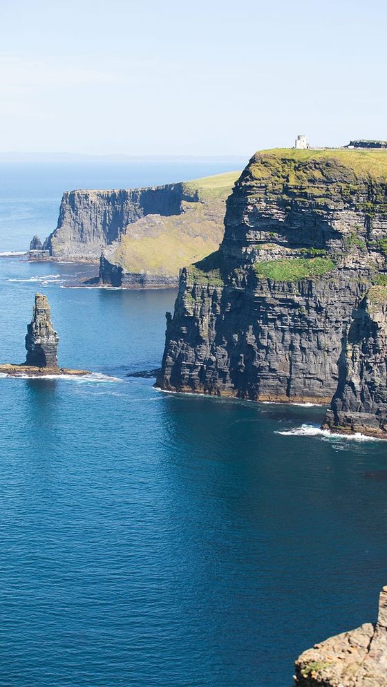 Cliffs of Moher with rugged rock formations and a lighthouse in the distance, surrounded by the blue Atlantic Ocean under a clear sky.
