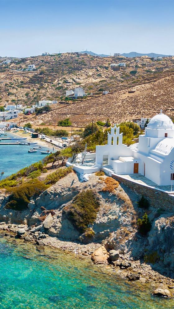 White church on a rocky cliff overlooking turquoise sea, with hills and buildings in the background under a clear blue sky.