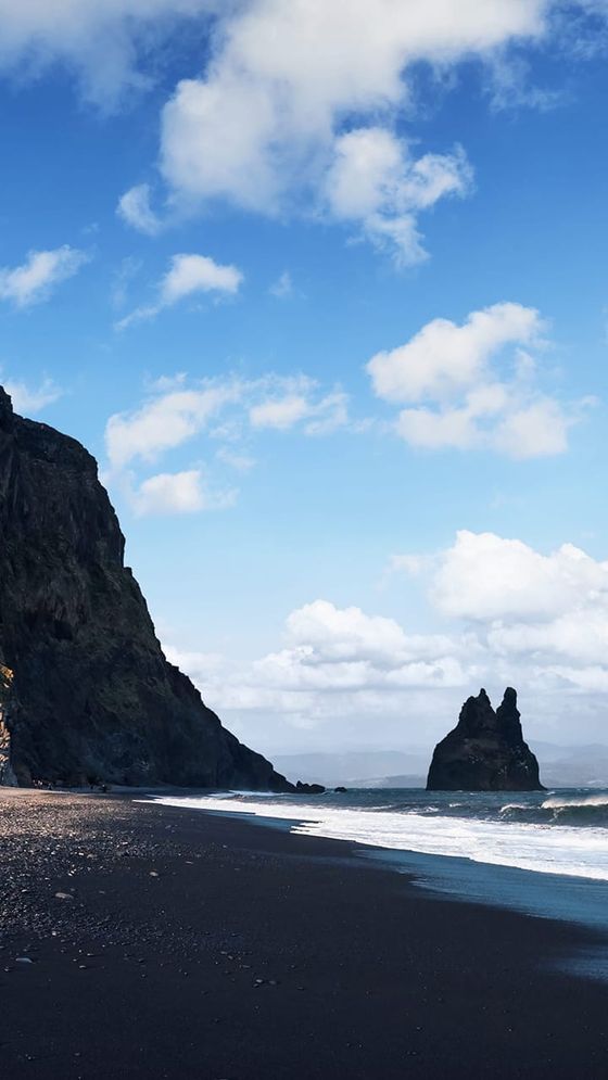 Black sand beach with rocky cliffs, ocean waves, and distant rock formations under a partly cloudy blue sky.