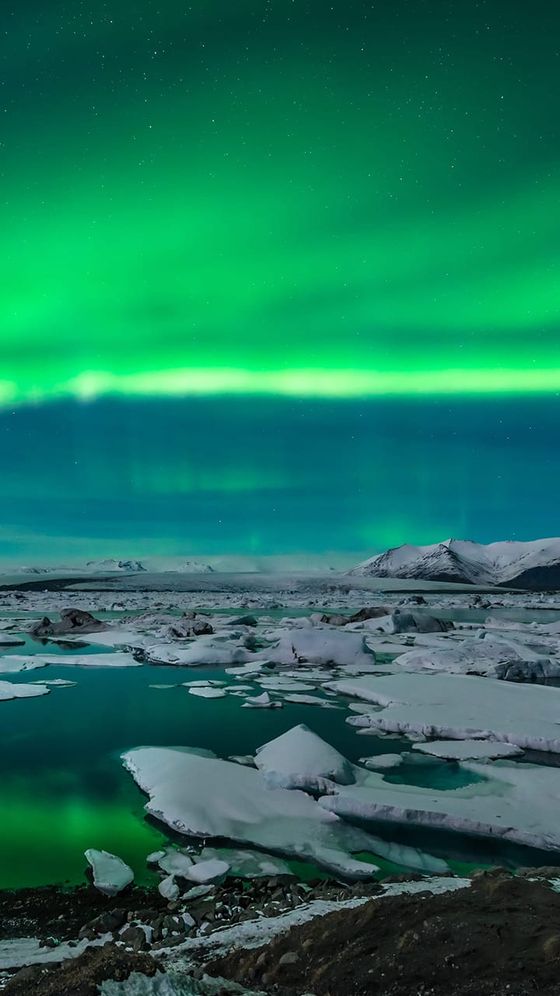 Northern lights display vibrant green hues over an icy landscape with scattered icebergs and distant snow-covered mountains.