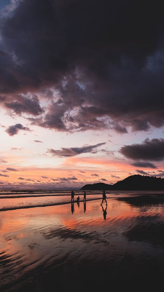 Silhouetted family walking on a reflective beach at sunset, with dramatic clouds and distant hills in the background.