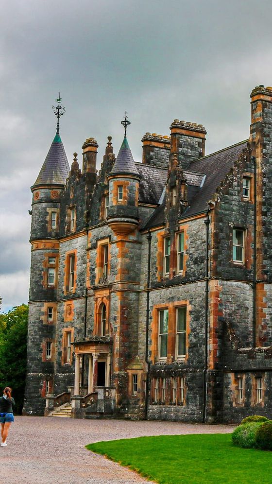 A person walks near a historic stone castle with conical towers and ornate details, set against a cloudy sky and surrounded by greenery.