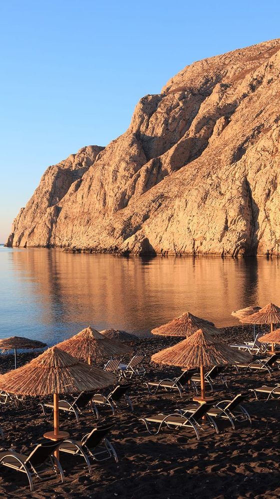Beach with straw umbrellas and lounge chairs on black sand, set against towering rocky cliffs by calm blue sea at sunset.