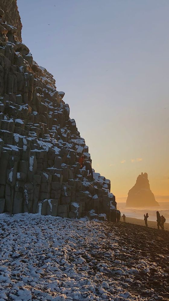 People exploring snow-dusted basalt columns by the ocean at sunset, with a rocky sea stack in the background.