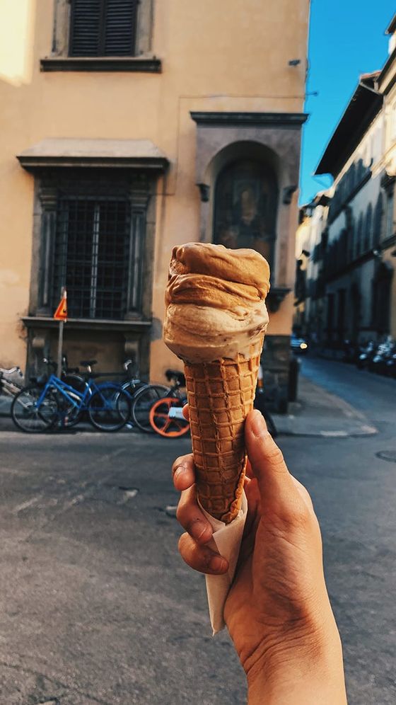 A hand holds a tall, swirled ice cream cone on a street with bicycles and historic buildings in the background.