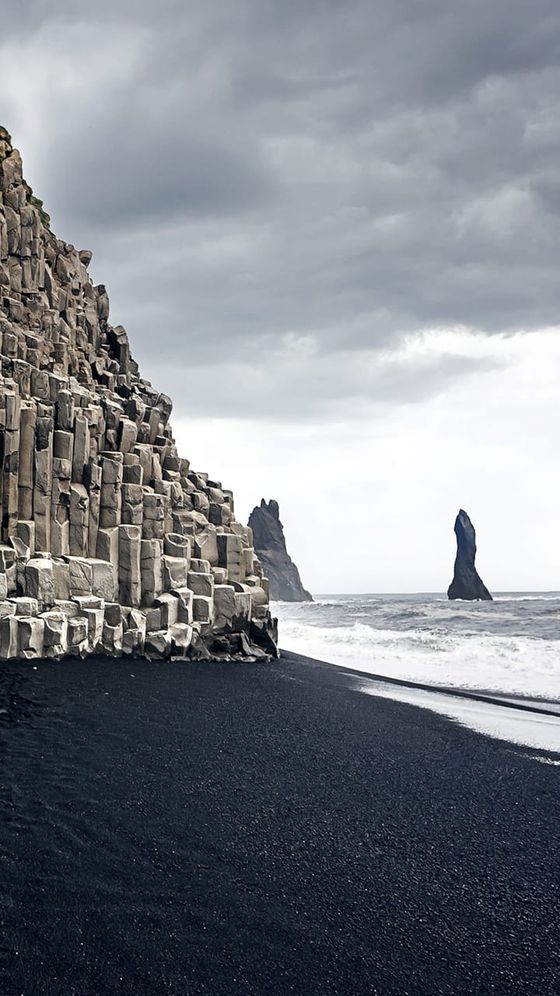 Dramatic rocky basalt columns on a black sand beach with rough seas and overcast skies, featuring sea stacks in the distance.