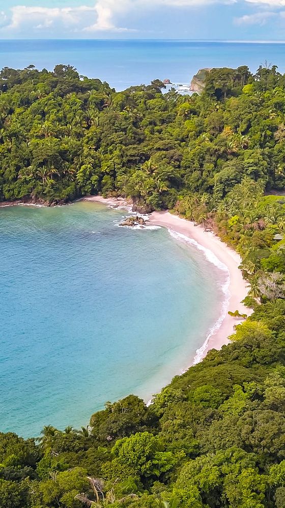 Lush green forest surrounding a secluded, crescent-shaped sandy beach with turquoise water and distant cliffs under a partly cloudy sky.