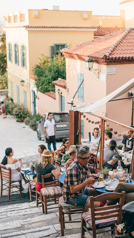 Outdoor café scene on a sunlit street with people dining at tables, surrounded by colorful buildings and greenery.
