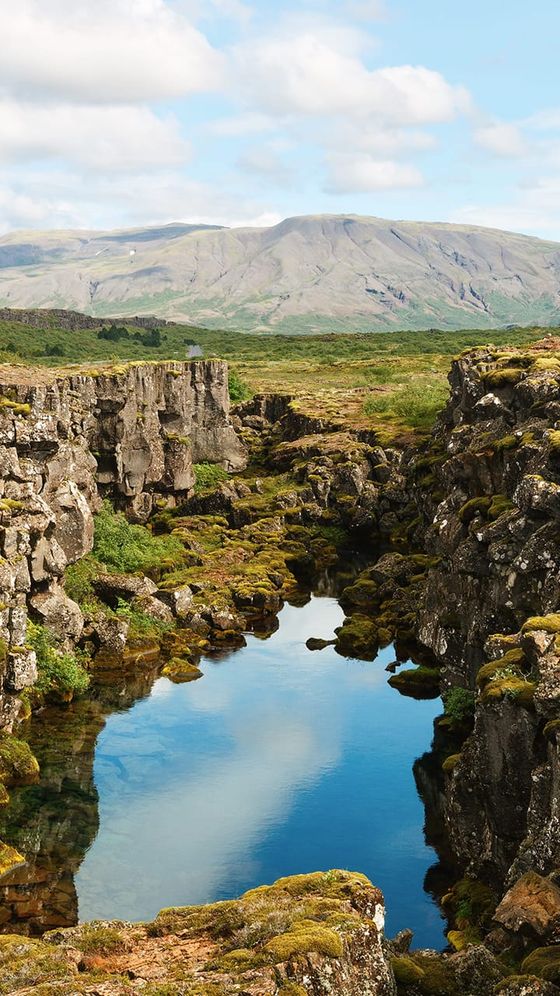 Rocky canyon with clear water reflecting the sky, surrounded by moss-covered cliffs and distant mountains under a partly cloudy sky.