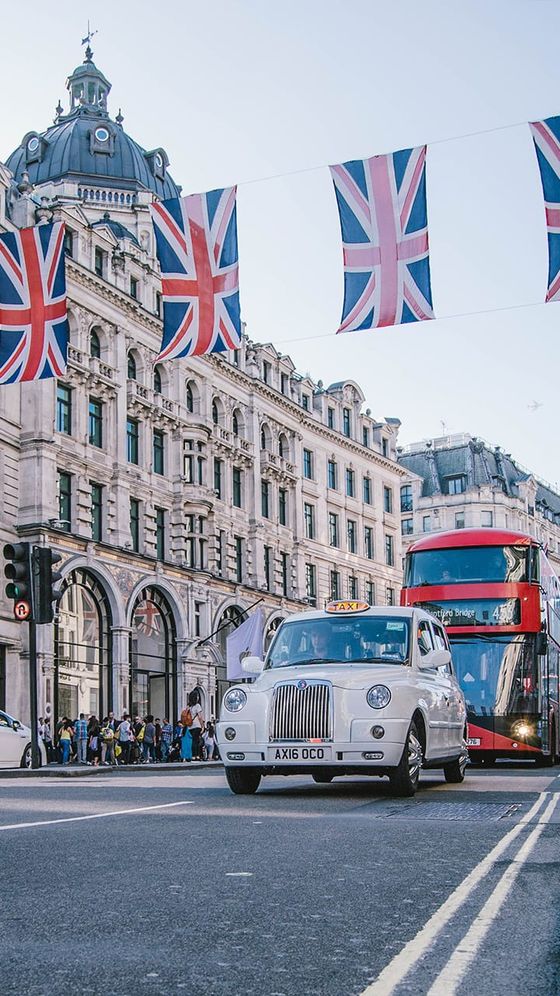 A classic white taxi and a red double-decker bus drive on a street adorned with Union Jack flags in front of historic buildings.