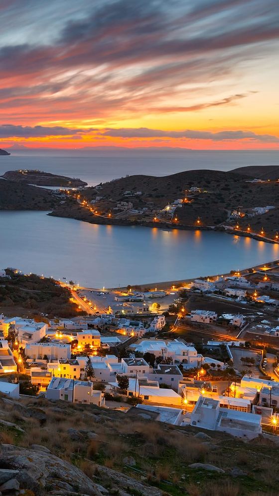 Sunset view over a coastal town with white buildings, a bay, and hilly landscape. Streetlights illuminate the scene as the sky glows with vibrant colors.