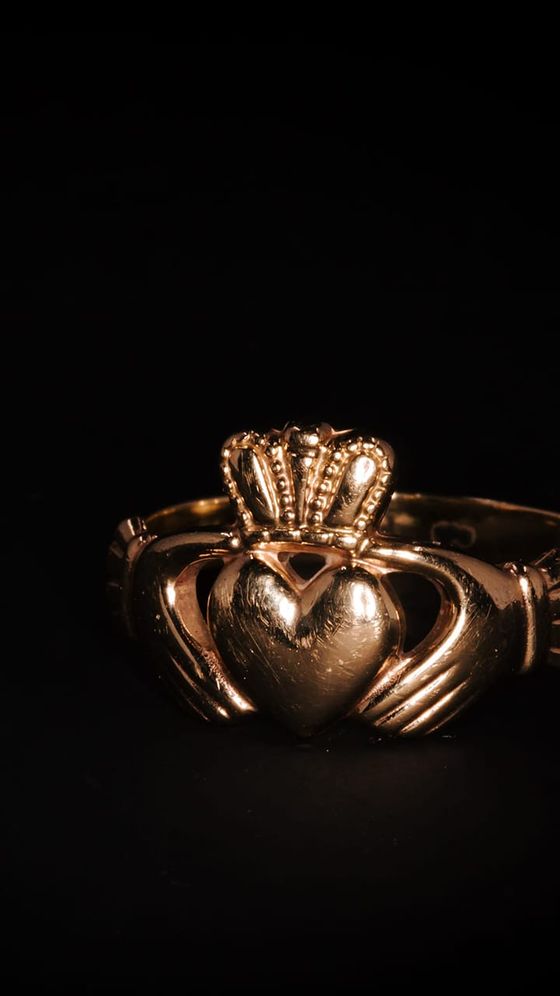 A gold Claddagh ring with hands holding a crowned heart, symbolizing love, loyalty, and friendship, set against a dark background.