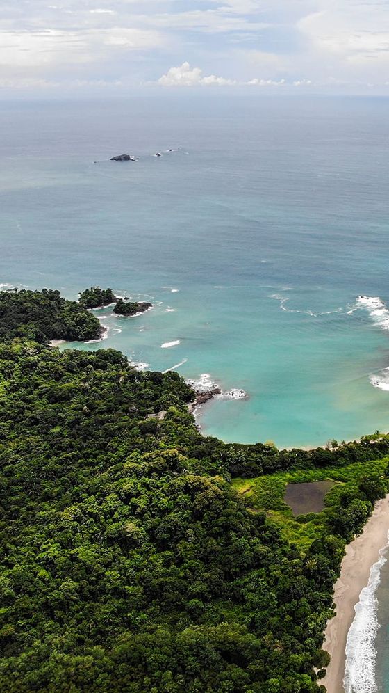 Aerial view of a lush green coastline meeting turquoise ocean, with rocky outcrops in the water and a sandy beach in the foreground.