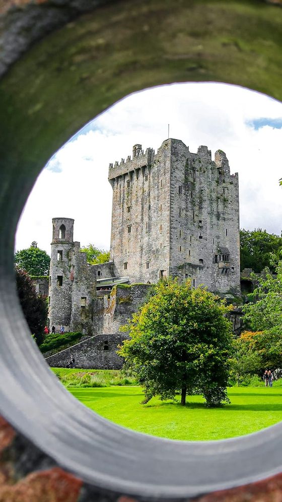 View of Blarney Castle through a circular stone frame, surrounded by lush greenery and a cloudy sky.