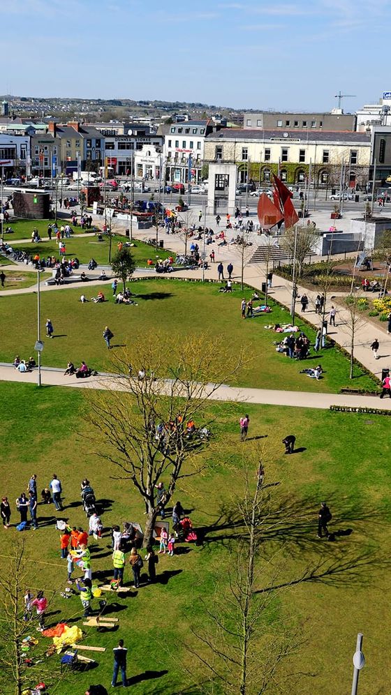Aerial view of a busy park with people relaxing on the grass and walking on paths, surrounded by buildings and a large sculpture.