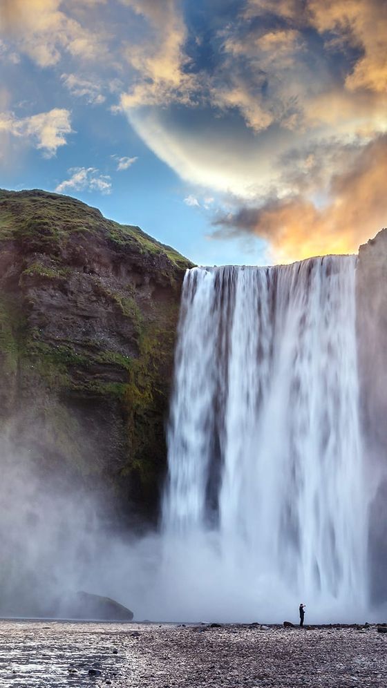 A person stands near the base of a large waterfall cascading over a rocky cliff, with a dramatic sky and mist surrounding the area.