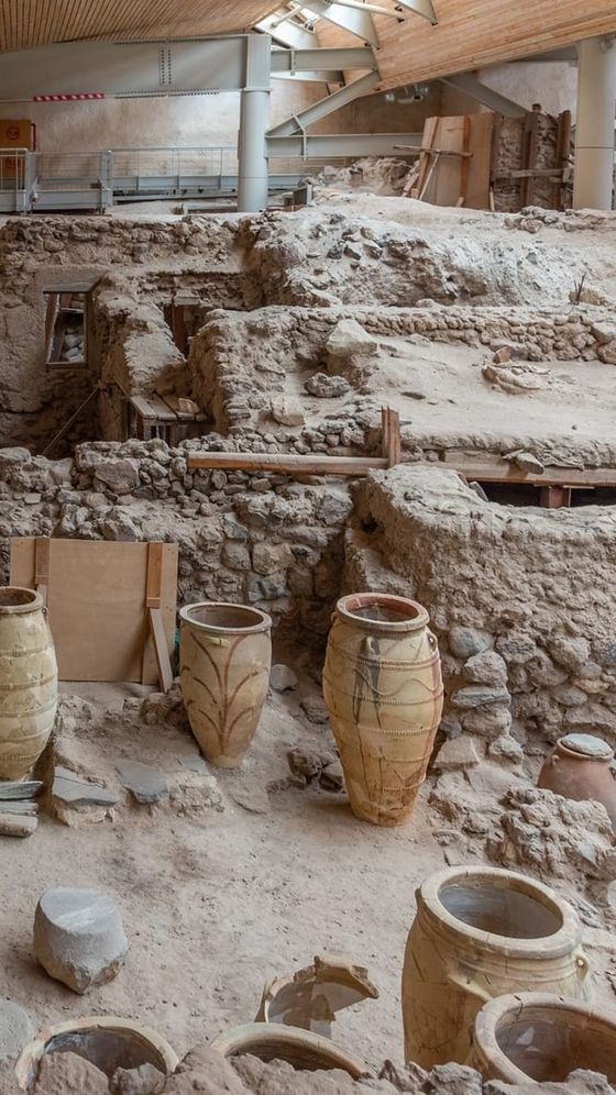 Ancient archaeological site with large pottery jars, stone walls, and excavated earth, under a modern roof structure.