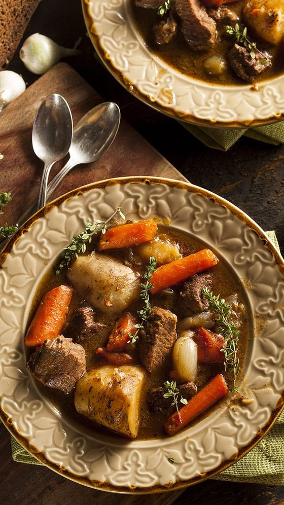 A bowl of hearty beef stew with chunks of beef, carrots, potatoes, onions, and herbs, set on a wooden table next to two spoons.