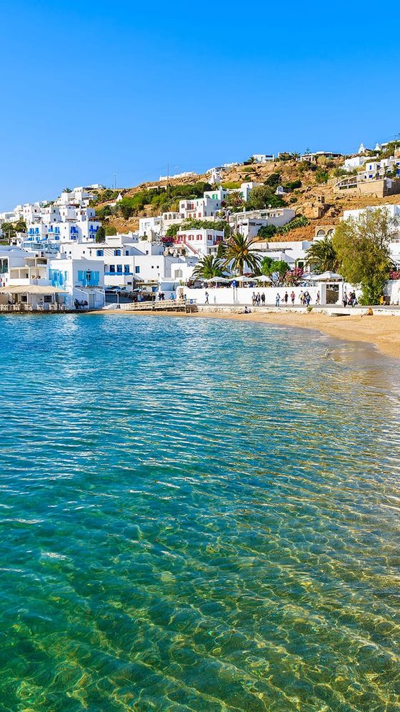 A clear turquoise beach with gentle waves, bordered by white buildings on a sunlit hillside under a bright blue sky.