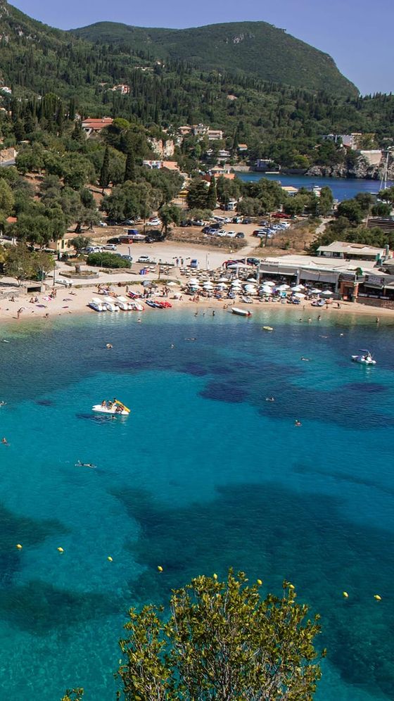 Aerial view of a beach with clear turquoise water, boats, sunbathers, and lush green hills in the background.