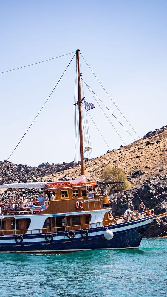 Sailing ship with wooden deck and people on board, cruising in turquoise waters near a rocky, arid coastline under a clear blue sky.
