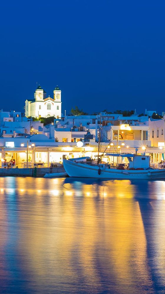 Seaside town at night with illuminated buildings, a small church in the background, and boats reflecting on calm water.