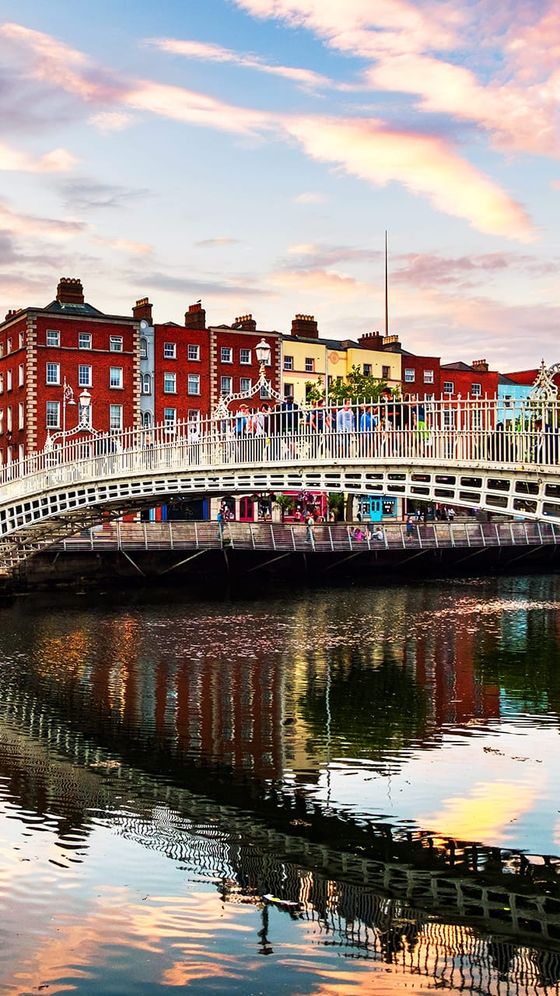 A picturesque view of Dublin's Ha'penny Bridge with colorful buildings in the background and reflections in the river below at sunset.