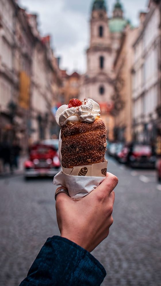 A hand holding a chimney cake topped with whipped cream and a strawberry, with a cobblestone street and historic buildings in the background.