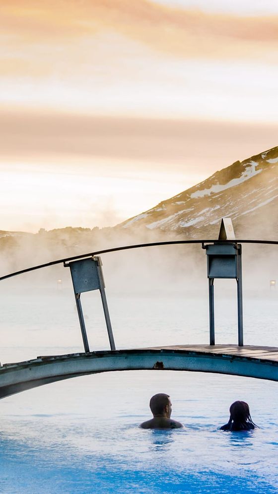 Two people relax in a steaming hot spring under a small bridge, with snow-capped mountains and a colorful sky in the background.