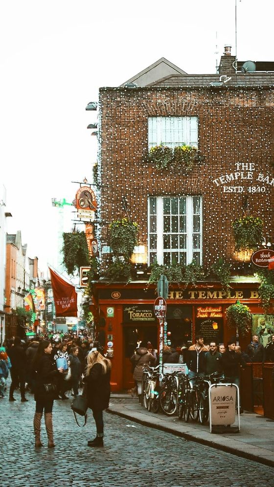 Street scene in front of The Temple Bar, Dublin, with people walking on cobblestone pavement, bicycles, and festive lights adorning the brick building.