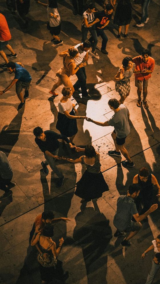 Aerial view of a lively group of people dancing on an outdoor stone floor, casting long shadows in warm lighting.