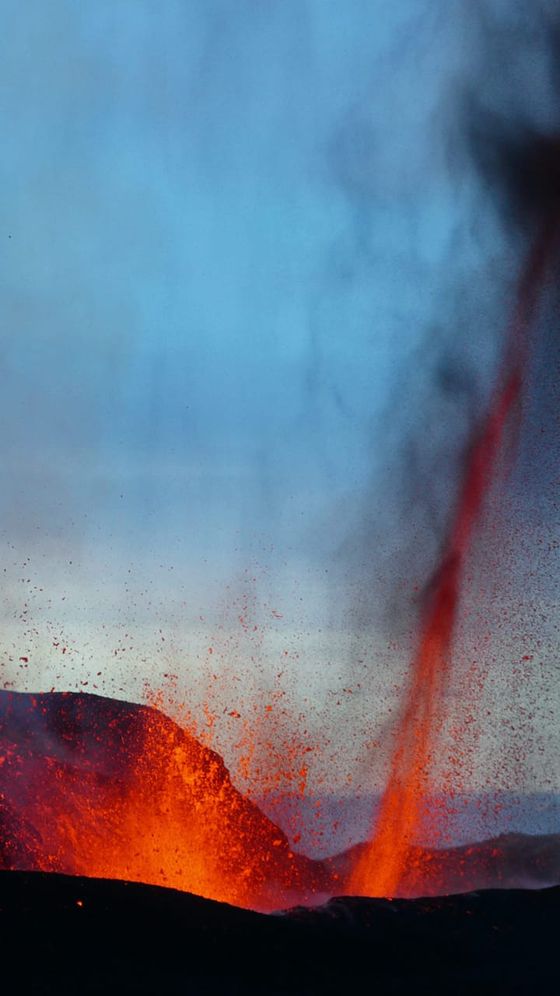Bright orange lava erupts from a volcanic crater, spewing into the sky against a twilight backdrop of deep blue and smoky grey.