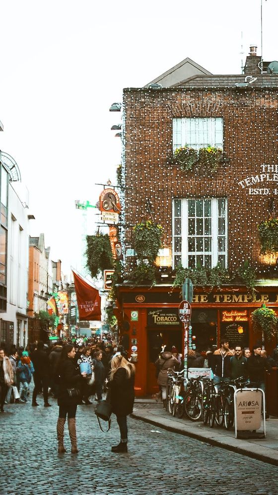 Cobblestone street in front of a lively pub adorned with string lights and flower boxes. People are gathered around, and bicycles are parked nearby.