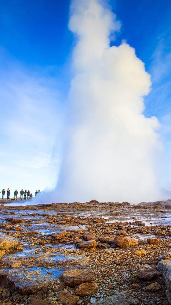 A powerful geyser erupts against a clear blue sky, with a group of people observing from a distance, standing on rocky terrain.