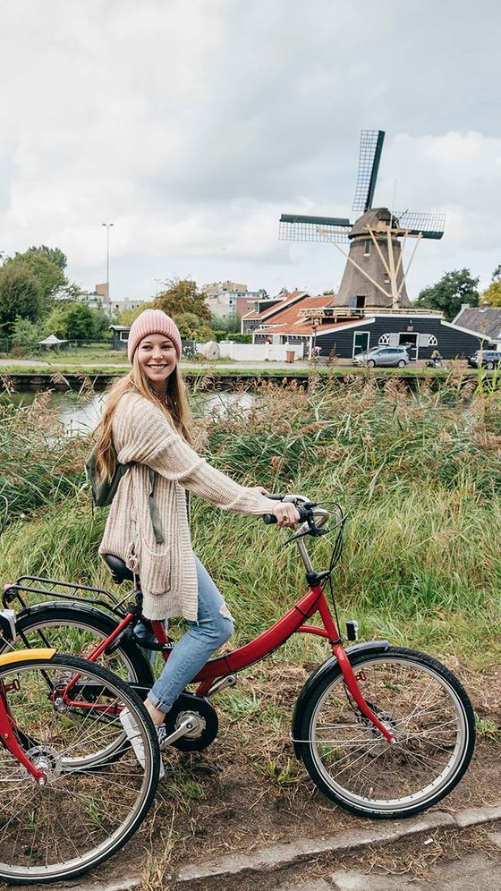Woman smiling on red bicycle near a grassy area with a windmill in the background, under a cloudy sky.