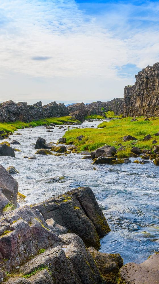 A tranquil river flows through rocky terrain with grassy banks under a partly cloudy sky. Majestic rock formations rise on either side.