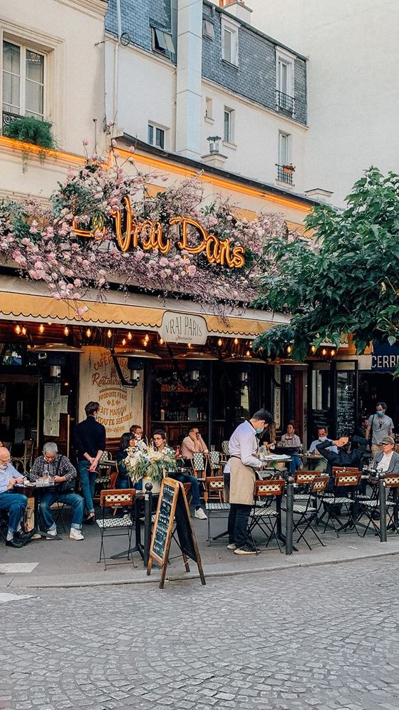 Street view of a Parisian café with outdoor seating, floral decor, patrons dining, and a waiter serving food on a cobblestone street.