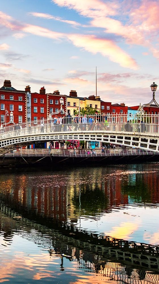 Historic arched bridge over a river with ornate railing, colorful buildings in the background, and a vibrant sky reflected in the water.