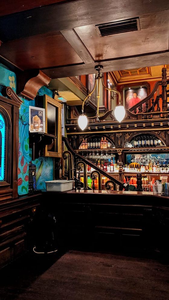 A dimly lit, ornate bar interior with wood paneling, decorative railings, colorful murals, and rows of liquor bottles on shelves.