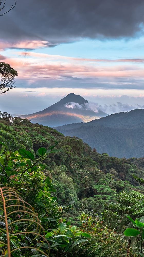 Scenic view of a distant volcano surrounded by lush green forest under a dramatic sky with clouds and patches of blue.