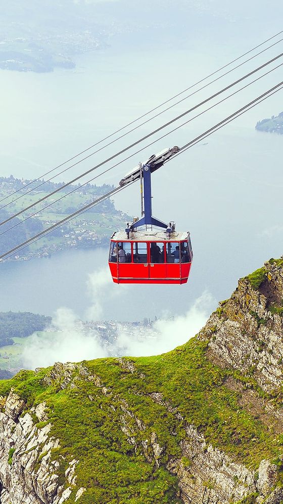 Red cable car above lush green mountains with a view of a lake and distant town below a cloudy sky.